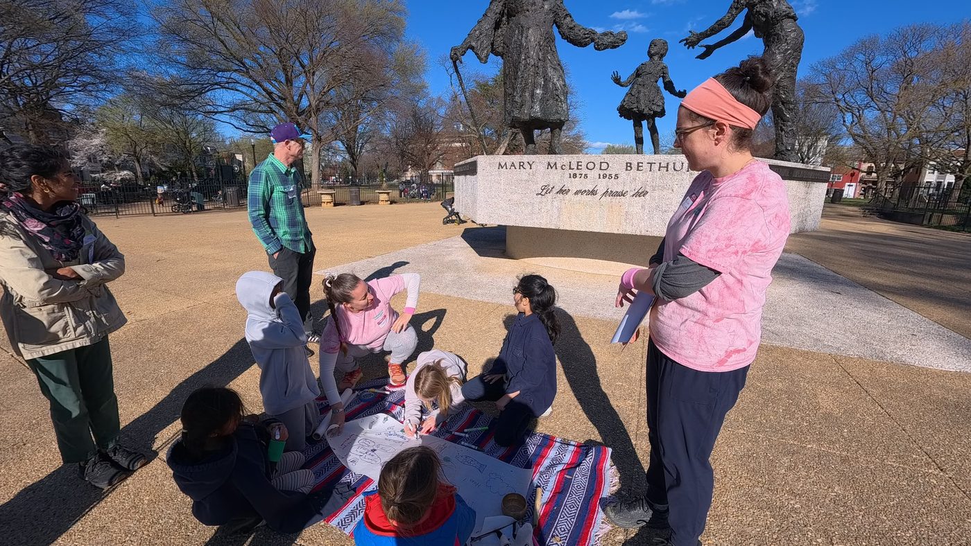 Kids draw together on a big sheet of paper on a blanket in front of the Mary McLeod Bethune memorial while adults look on