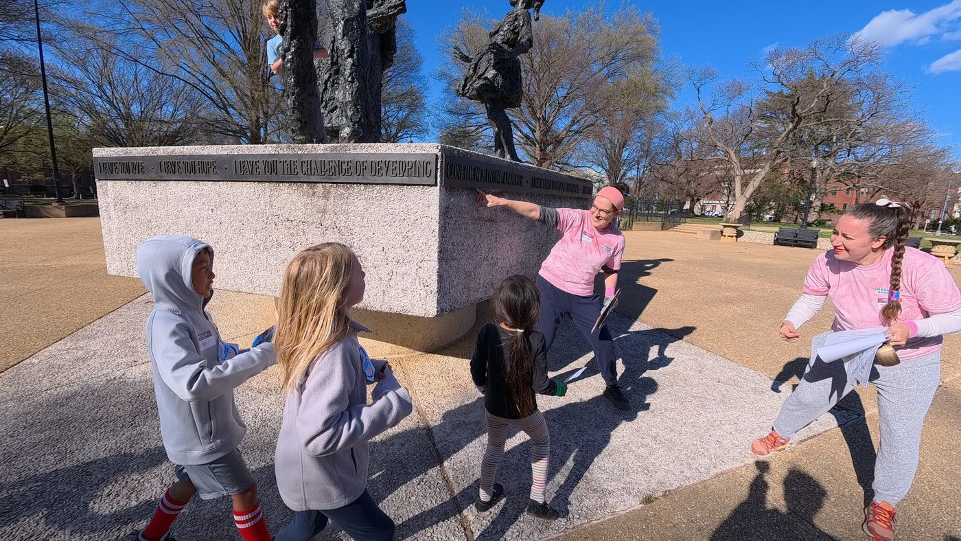 A facilitator in a pink Imagination Bootcamp shirt points out an inscription on the Mary McLeod Bethune memorial as kids gather round