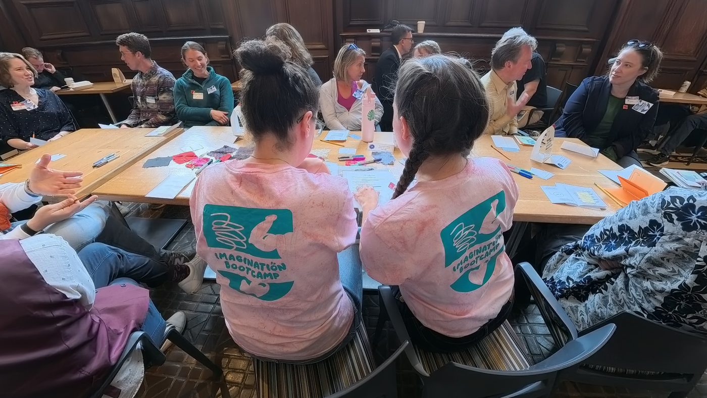 Isabella and Marina, in matching pink Imagination Bootcamp shirts, sit side by side at a long table of participants