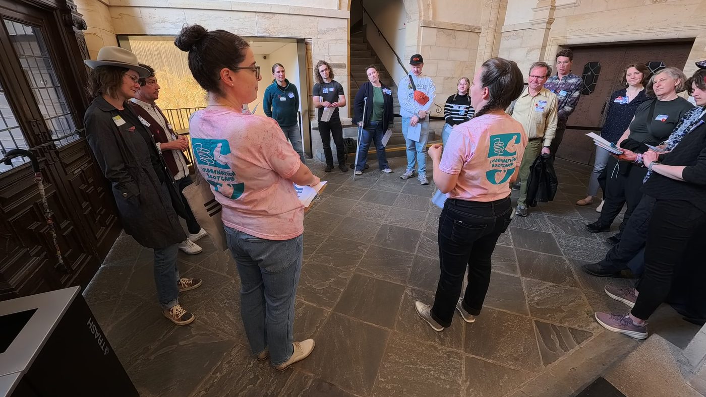 Adults stand in a circle in the Folger's stone entry hall with two facilitators in pink Imagination Bootcamp shirts