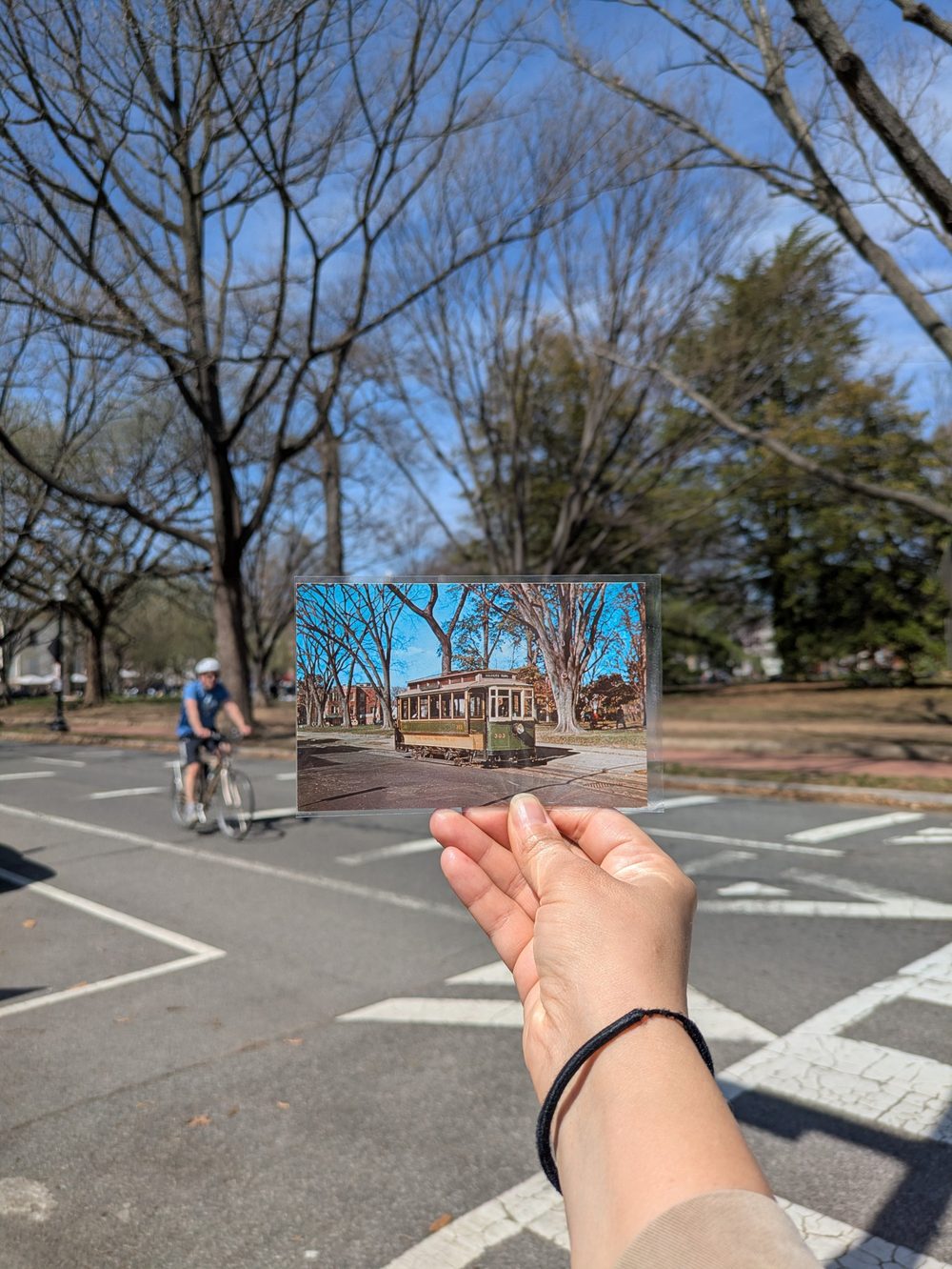 A hand holds up an old postcard of a streetcar at the same Lincoln Park corner where a cyclist now rides by
